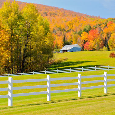 Daytime view of a barn nestled against a mountain covered in rich fall foliage with fields and a three-bar horse fence in the foreground