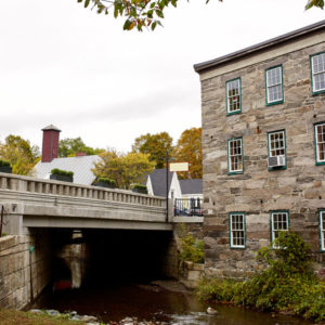Daytime view of a bridge over a creek next to a stone building in Vermont.
